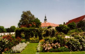 Historischer Garten mit Blumen und Bäumen, im Hintergrund ein Gebäude mit Turm.