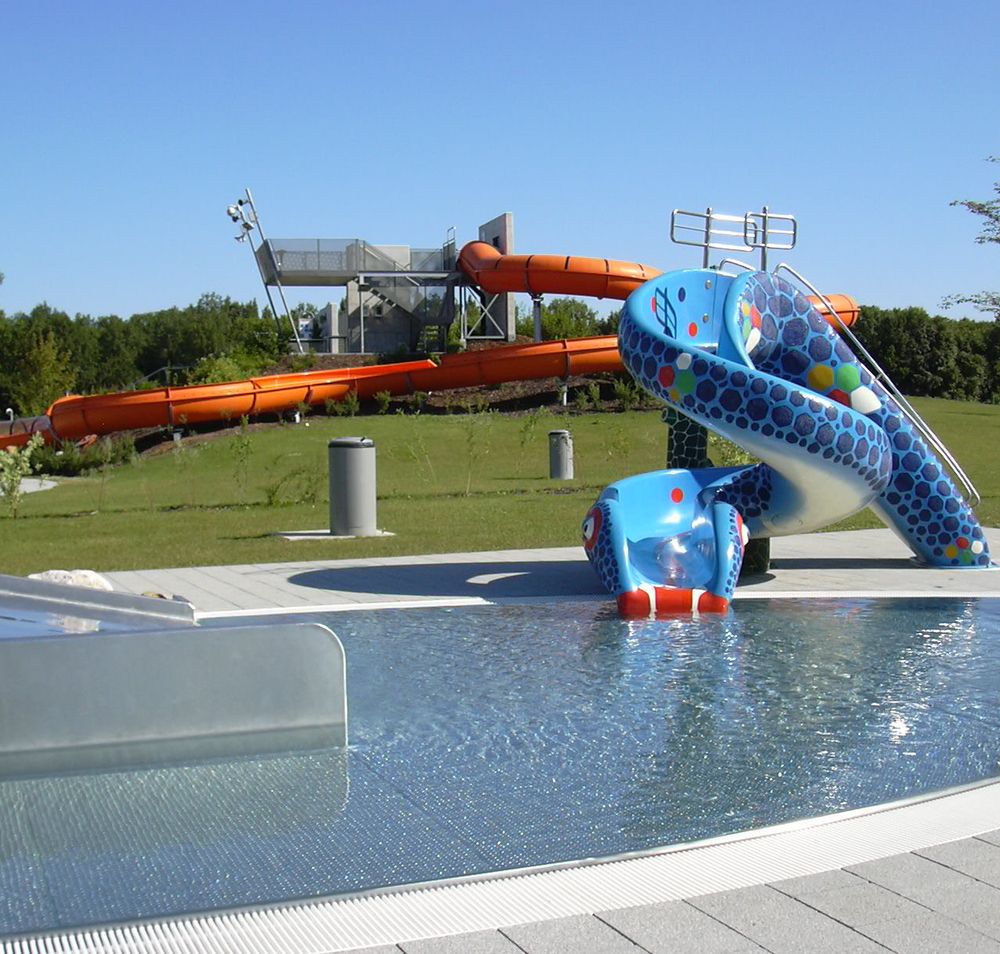 Blick auf einen Aquapark mit blauer Kinderrutsche und großer oranger Wasserrutsche im Hintergrund.
