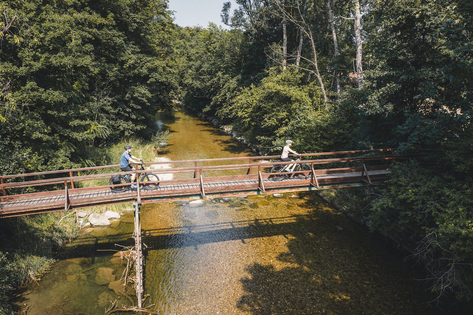 Zwei Personen fahren mit ihren Rädern über eine Holzbrücke über die Erlauf. Links und rechts neben dem Fluss dichte Pflanzen.