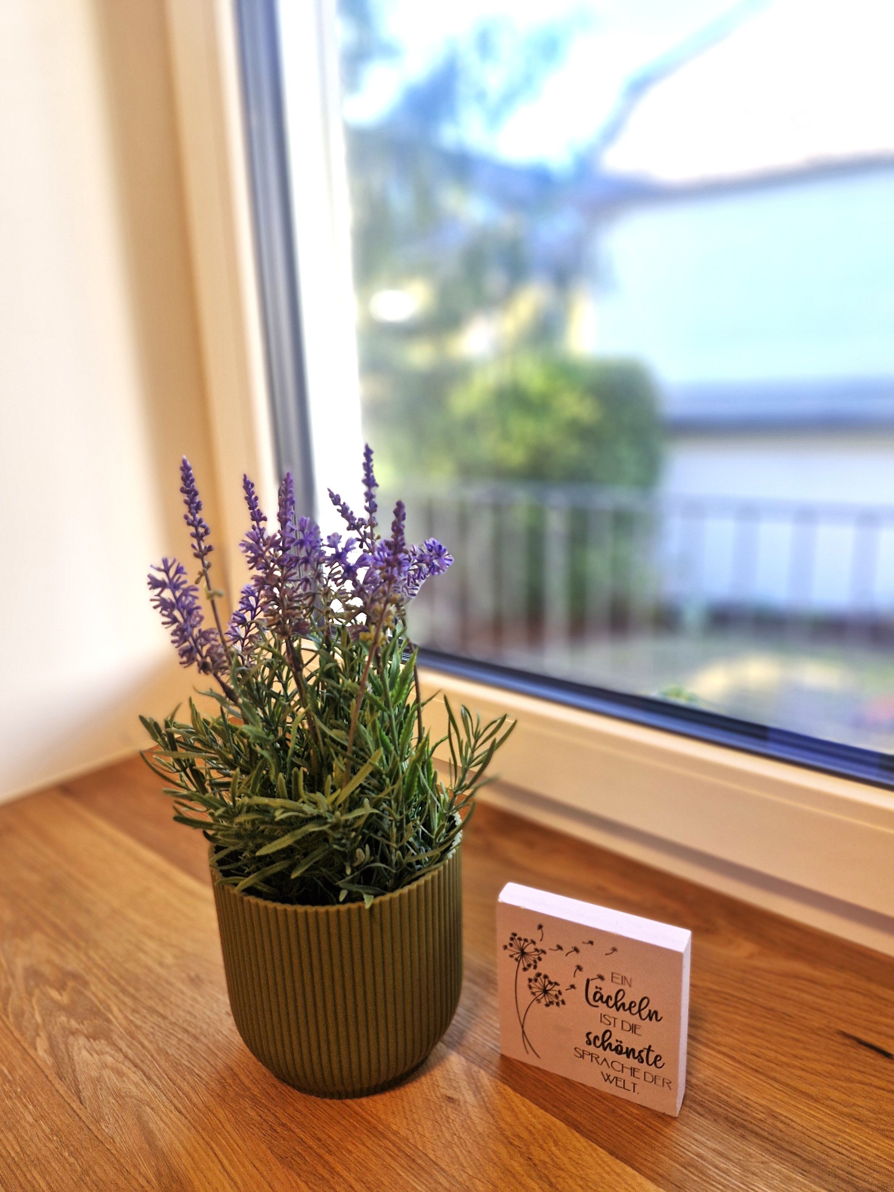 Ein Lavendeltopf und ein Schild mit einem Spruch stehen auf einer Fensterbank aus Holz.