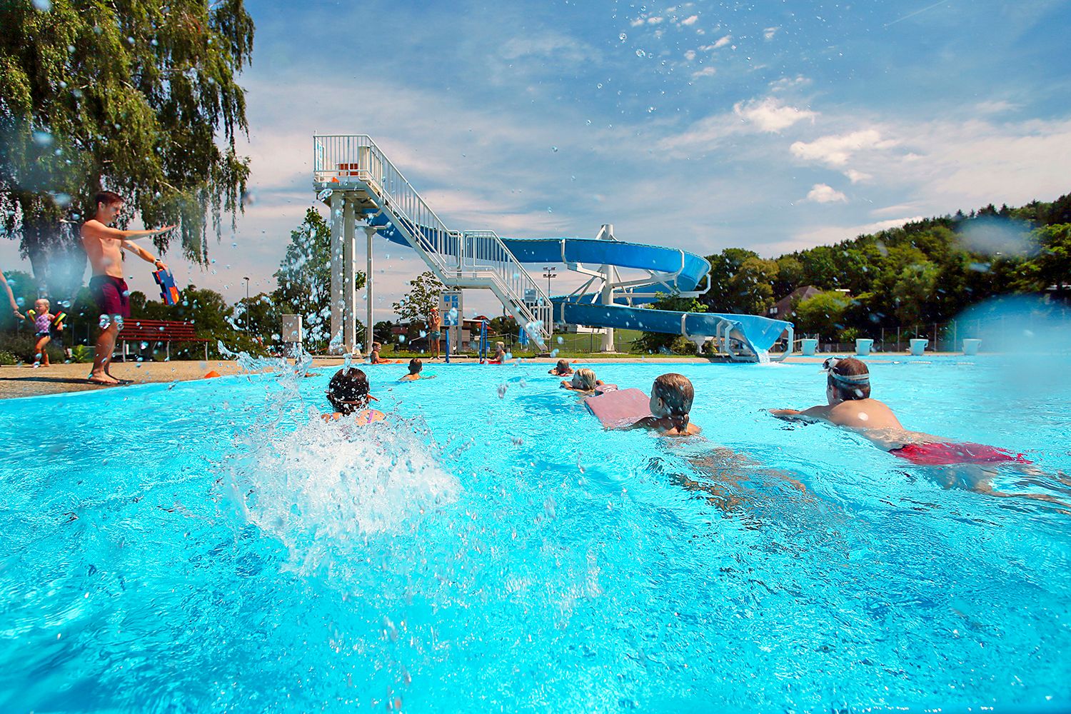 Kinder schwimmen im Freibad Neuhofen an der Ybbs mit einer großen Wasserrutsche im Hintergrund.