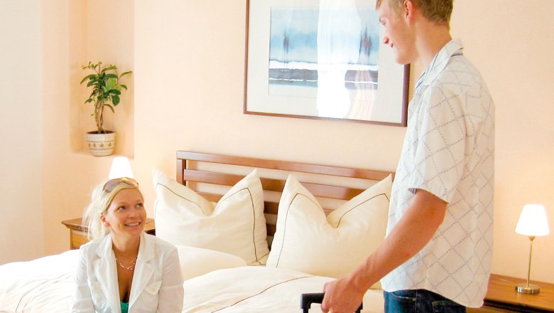 Ein Mann und eine Frau in einem Hotelzimmer, die Frau sitzt auf dem Bett, der Mann steht mit einem Koffer.