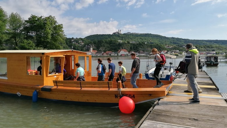 Menschen steigen auf ein Holzboot an einem Steg ein, im Hintergrund eine h&uuml;gelige Landschaft mit Geb&auml;uden.