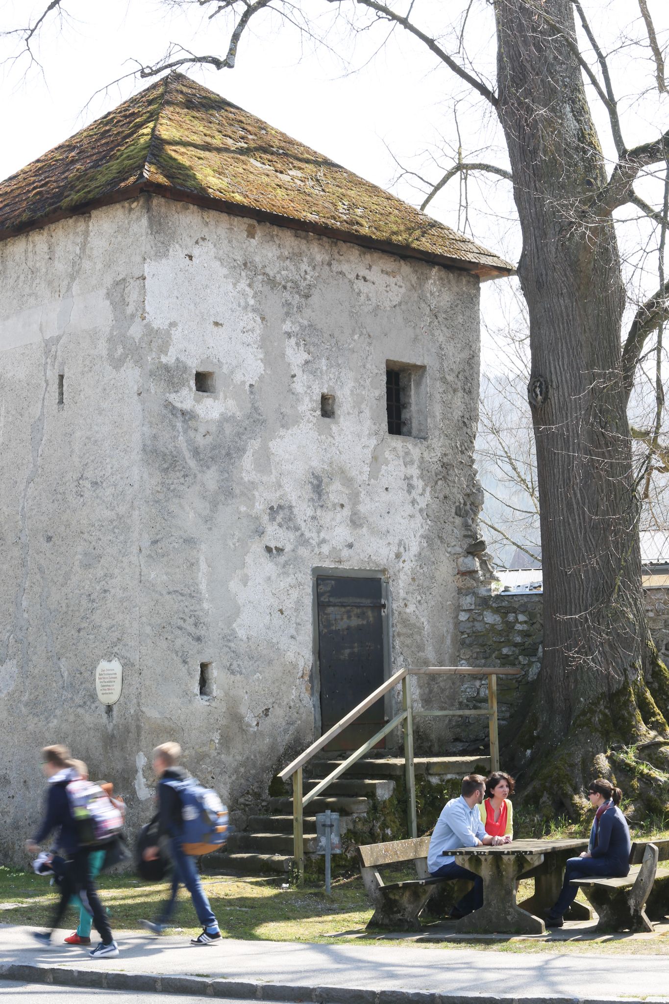 Ein alter Turm mit moosbedecktem Dach, daneben ein Baum. Drei Personen sitzen an einem Tisch, während zwei Personen vorbeigehen.