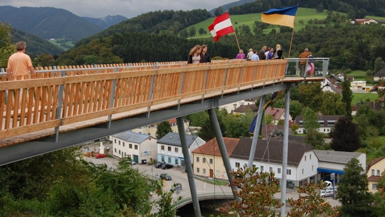 Menschen auf einer Holzplattform mit österreichischer und ukrainischer Flagge, im Hintergrund eine ländliche Landschaft.