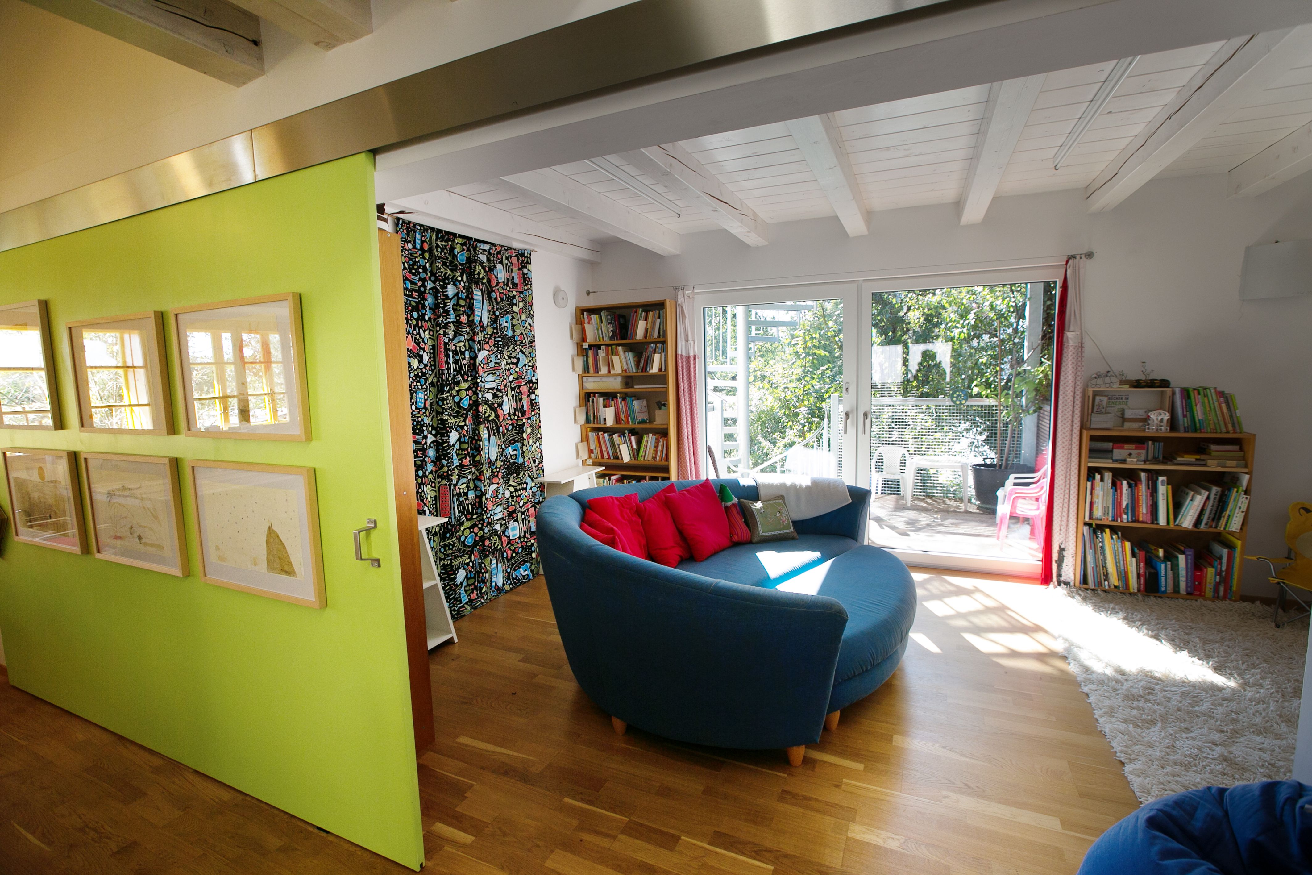 Innenansicht eines modernen Wohnzimmers mit runder blauer Couch, Holzboden, Bücherregalen und großen Fenstern mit Blick auf einen Garten.