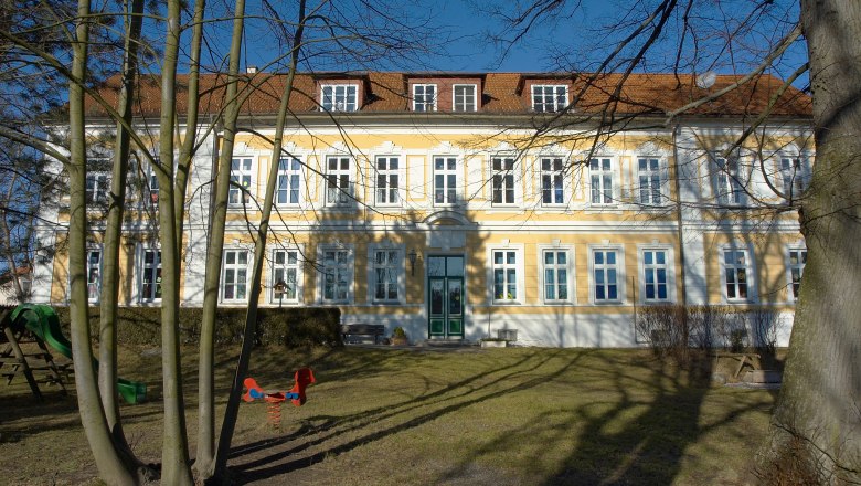 Gelbes Gebäude mit weißen Fensterrahmen und rotem Dach, umgeben von Bäumen und Spielplatz.