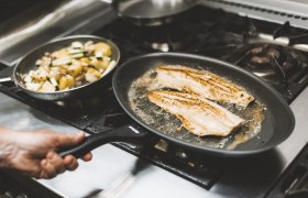 Two fish fillets fry in a pan on a stove, with a pan of vegetables next to them.