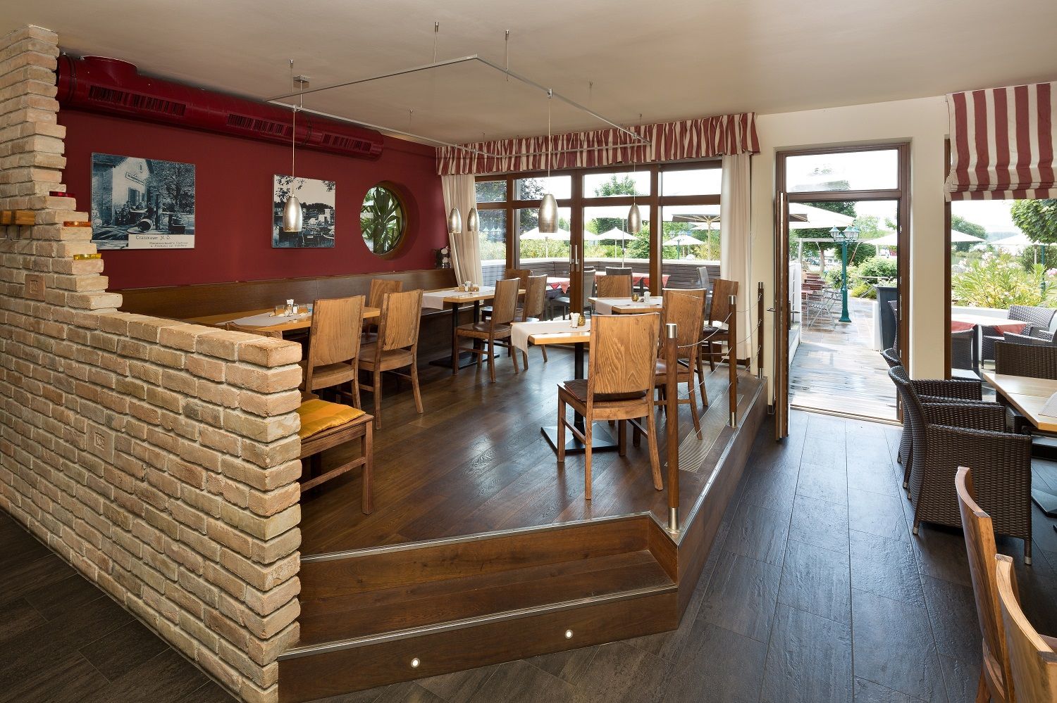 Ein gemütliches Restaurant mit Holzmöbeln, roten Wänden und großen Fenstern, die auf eine Terrasse führen.