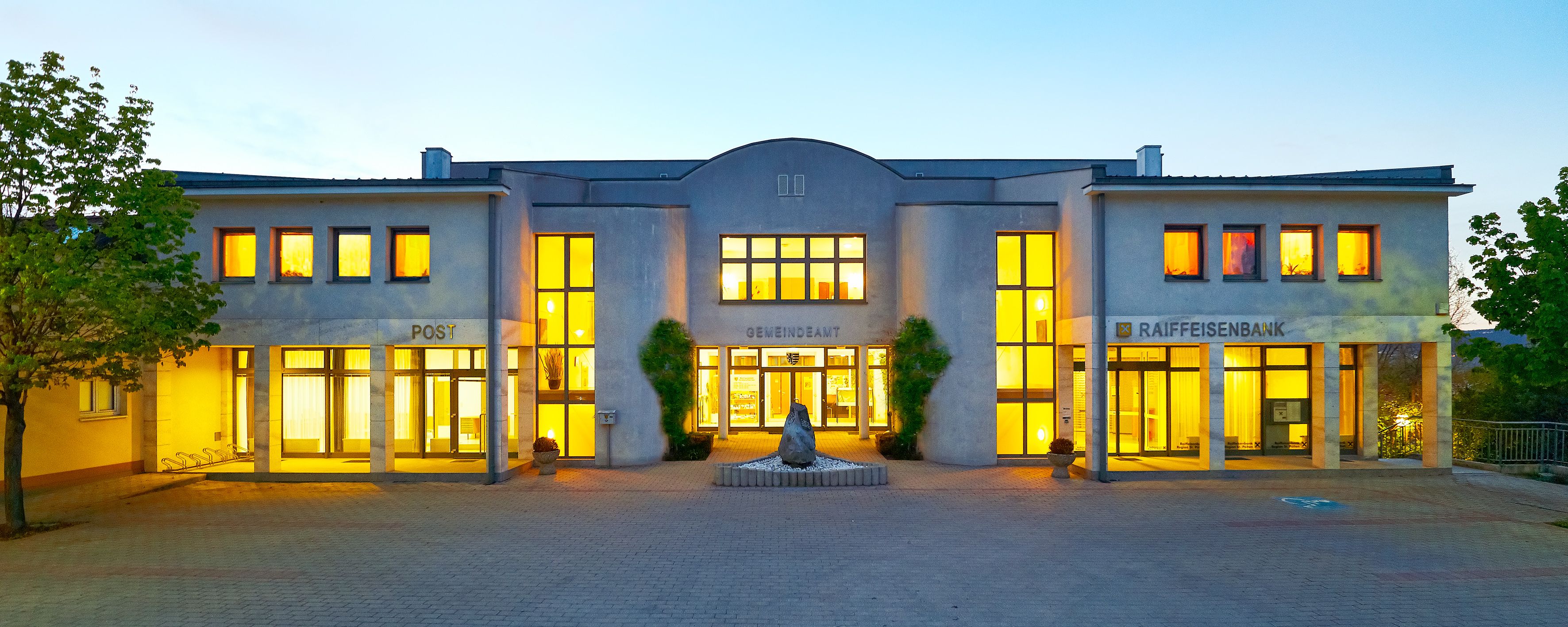 Beleuchtetes Gebäude mit Aufschrift 'Gemeindeamt', 'Post' und 'Raiffeisenbank'.