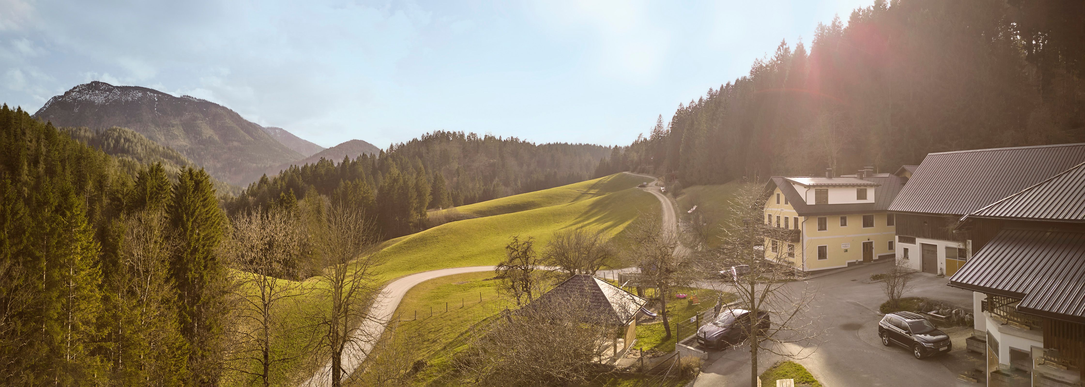 Panorama eines Bauernhofs in einer hügeligen Landschaft mit Wald und Bergen im Hintergrund.