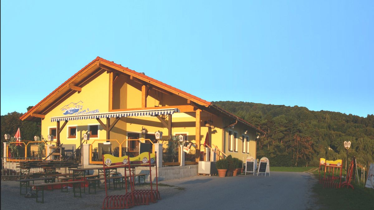Ein gelbes Gebäude mit Terrasse und Biergartenmöbeln, umgeben von grünen Hügeln unter klarem Himmel.