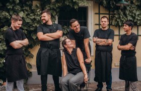 Eine Gruppe von sechs Personen in schwarzer Arbeitskleidung steht und sitzt lächelnd vor einer mit Weinreben bewachsenen Wand.