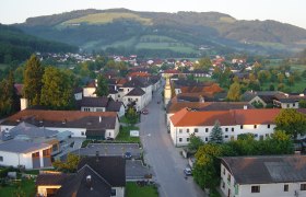 Luftaufnahme eines Dorfes mit roten D&auml;chern und gr&uuml;ner Landschaft im Hintergrund.