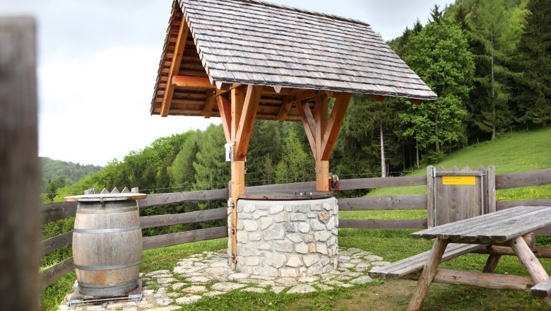 Ein traditioneller Brunnen mit Holzdach und Steinmauer, daneben ein Holzfass und eine Picknickbank, umgeben von grüner Landschaft.