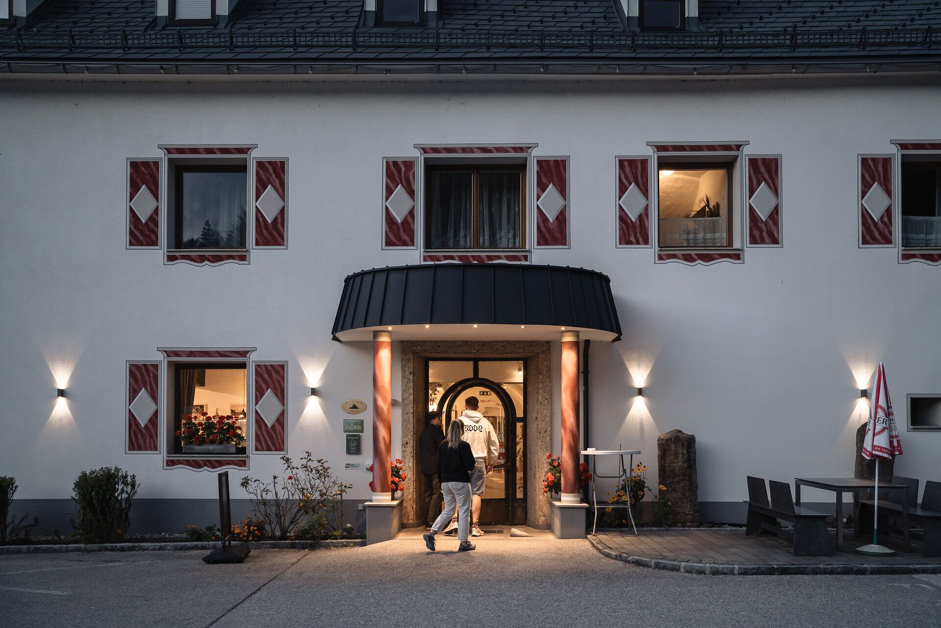 Der Eingangsbereich des Landhotels und Restaurants Zellerhof in Lunz am See bei Abenddämmerung. Drei Personen betreten das Gebäude durch eine einladend beleuchtete Tür, eingerahmt von dekorativen Fensterläden. Die Beleuchtung und das äußere Erscheinungsbild strahlen eine warme, gastfreundliche Atmosphäre aus.