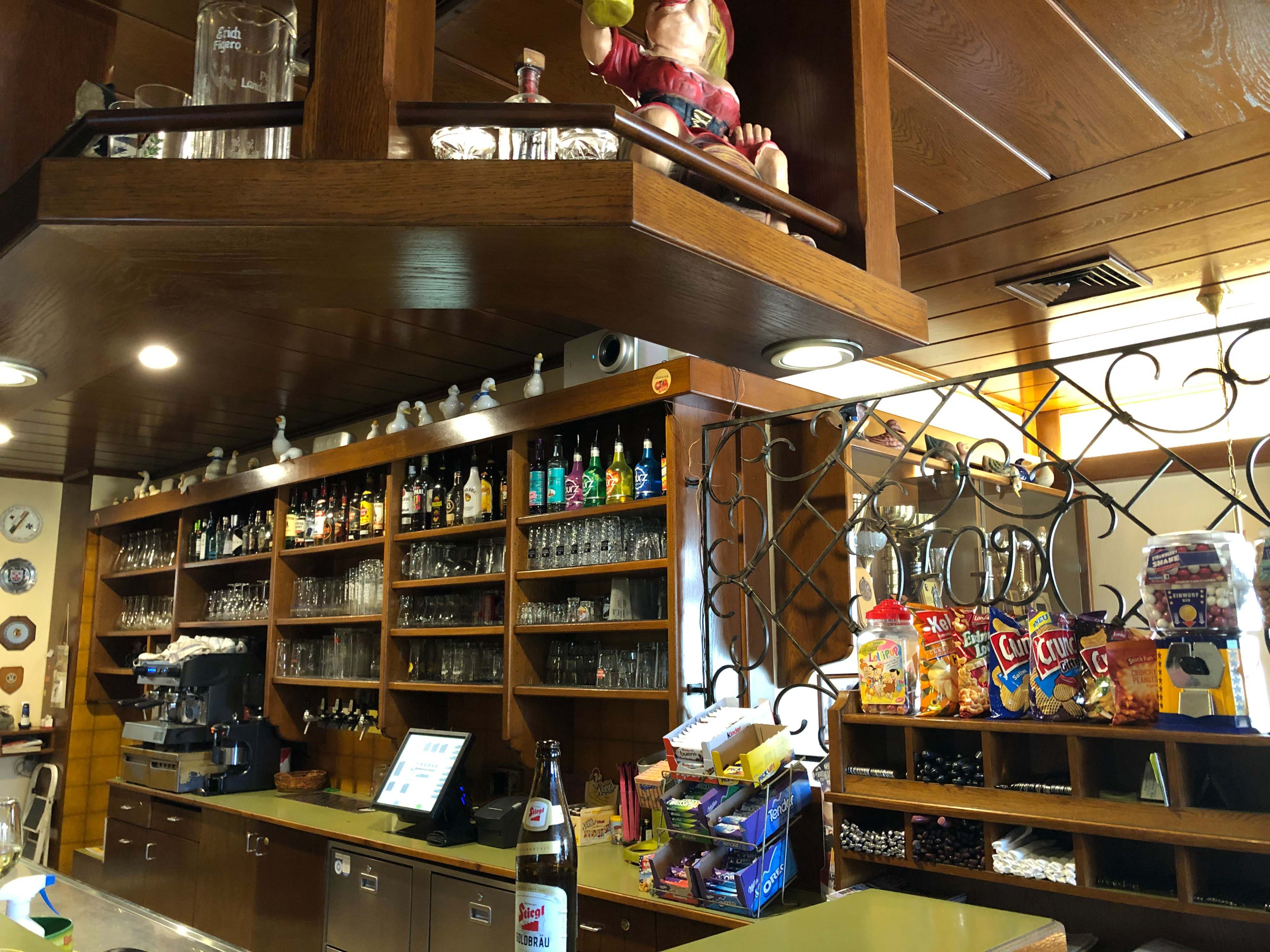 Innenansicht einer Bar mit Holzregalen, Gläsern, Flaschen und Snacks.