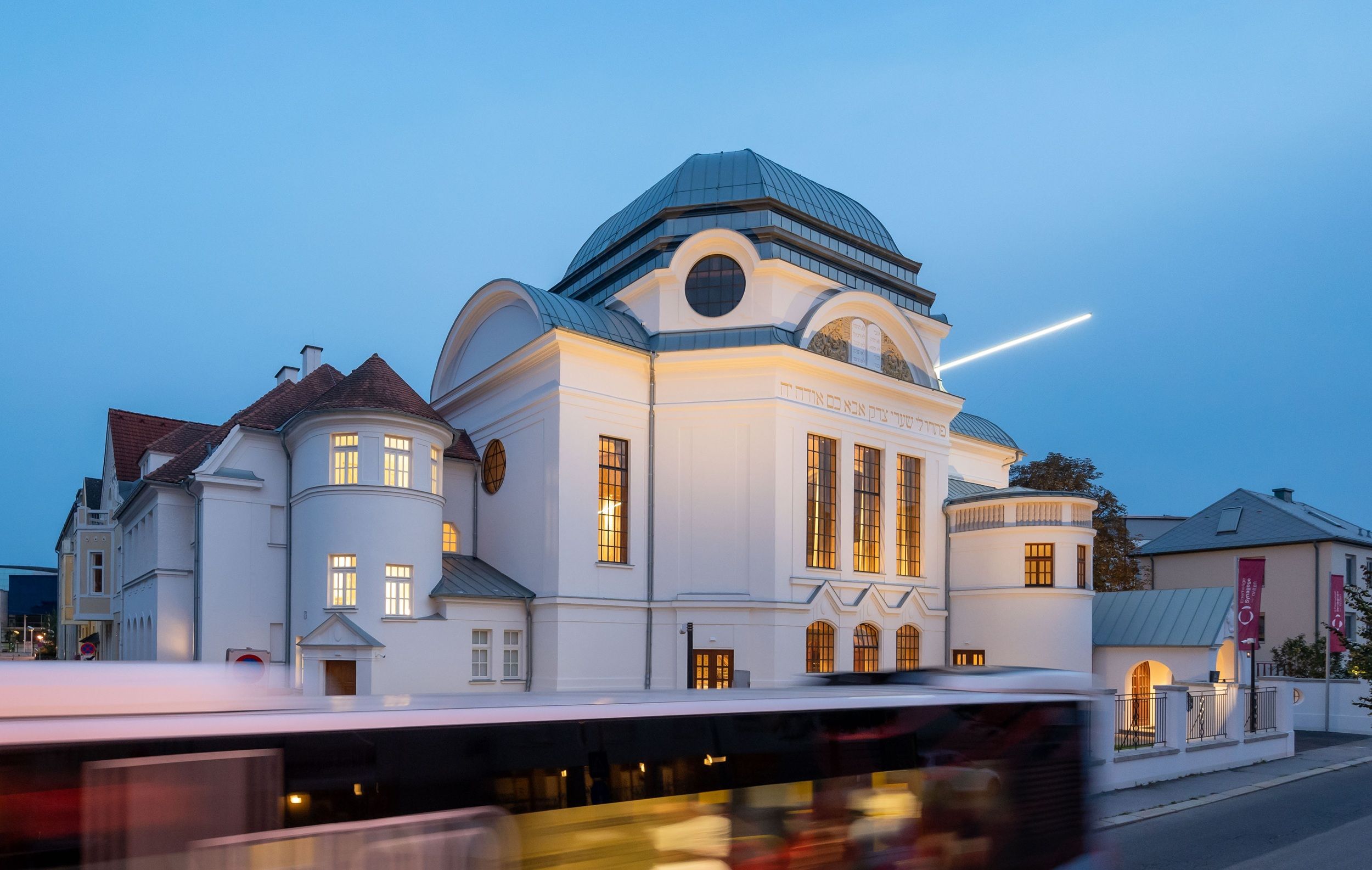 Ehemalige Synagoge in St. Pölten bei Dämmerung mit beleuchteten Fenstern und vorbeifahrendem Bus.