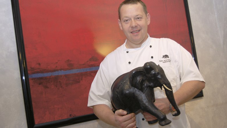 Ein Koch in weißer Uniform hält eine schwarze Elefantenfigur vor einem roten Gemälde.