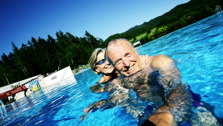An elderly couple smiling as they swim in an outdoor pool with a forest in the background.