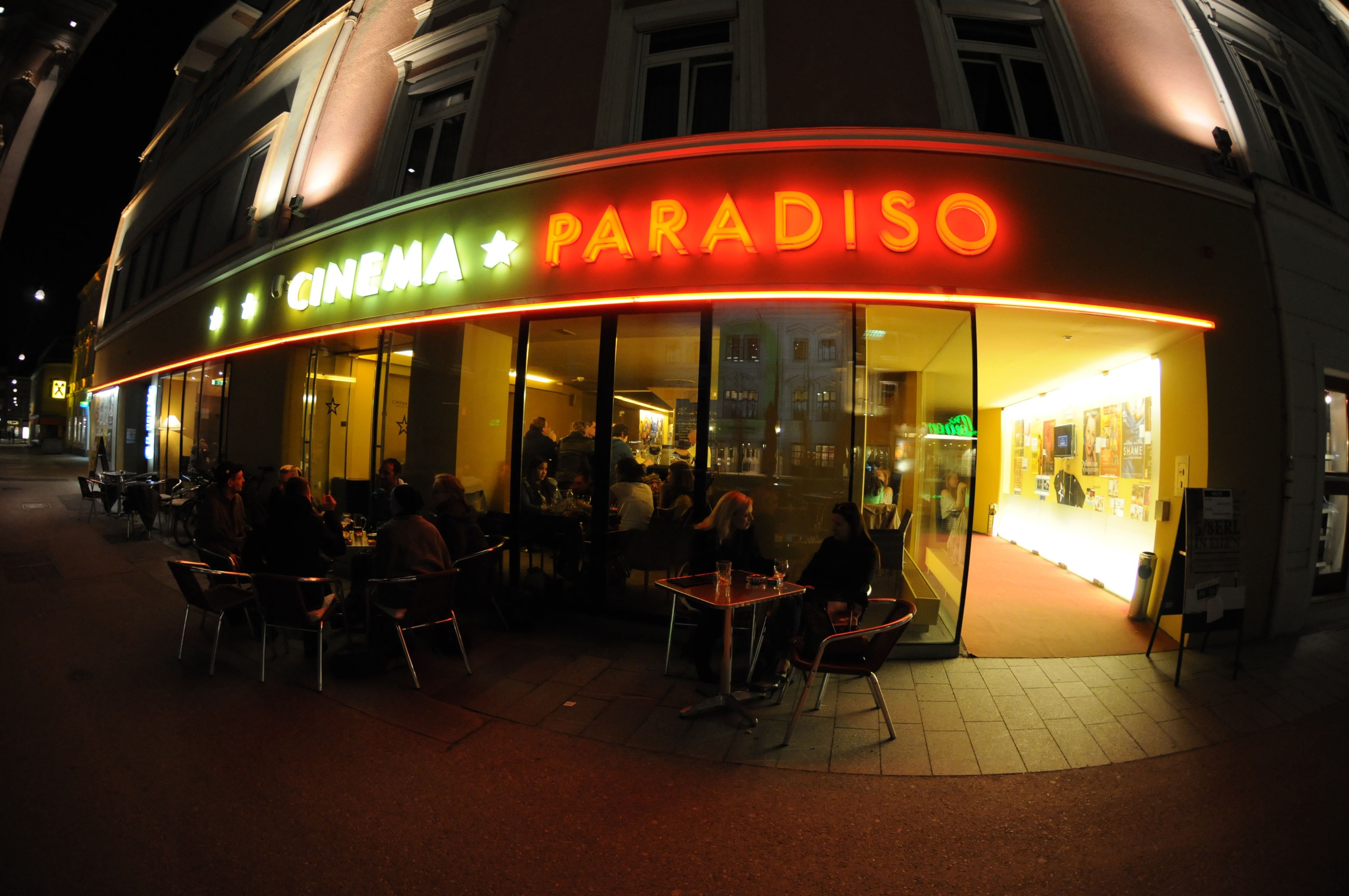 Außenansicht eines Kinos namens Cinema Paradiso bei Nacht mit beleuchtetem Eingang und Menschen an Tischen.