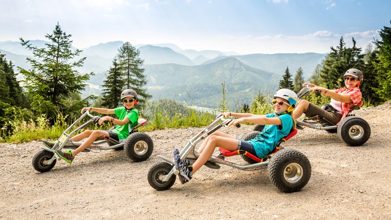 Drei Personen fahren auf Mountaincarts einen Berg hinunter, umgeben von einer Bergkulisse.