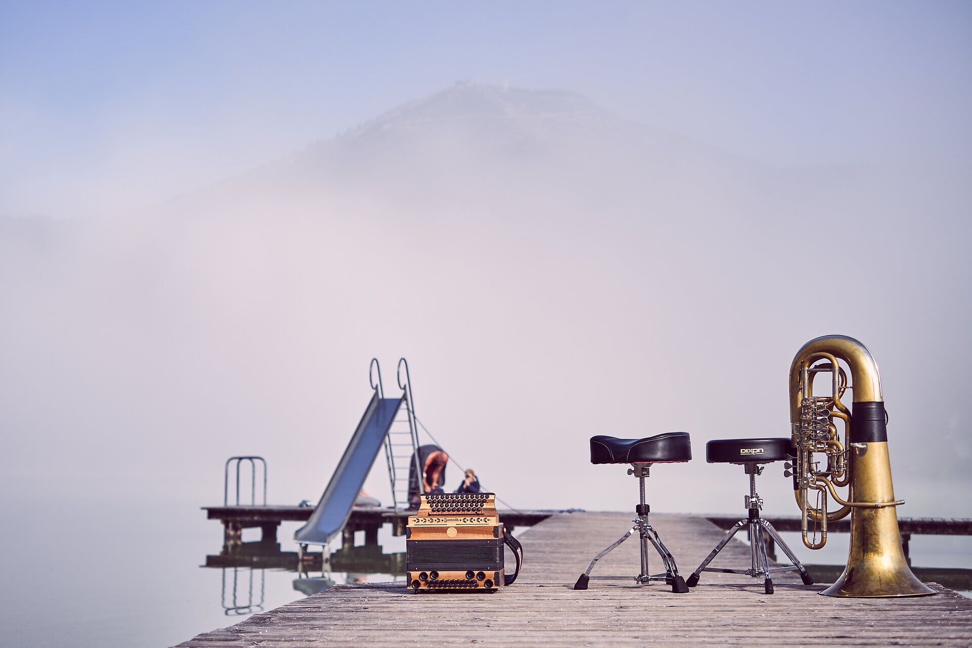 Ein sanfter Nebel umhüllt den ruhigen Erlaufsee, während die Klänge von Musikinstrumenten die Luft erfüllen. Auf dem Holzsteg, der ins Wasser führt, laden die harmonischen Töne dazu ein, die Seele baumeln zu lassen und die Schönheit der Natur zu genießen.