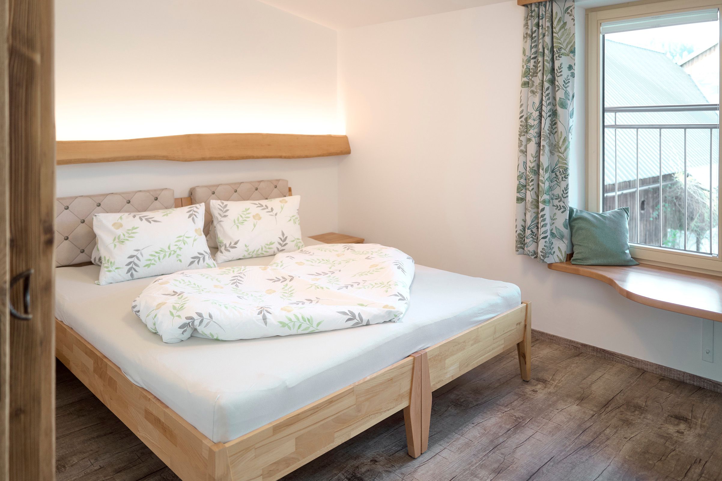 Ein gemütliches Schlafzimmer mit einem Holzbett, dekoriert mit Pflanzenmotiven auf Bettwäsche und Vorhängen. Ein Fenster mit Sitzbank bietet Ausblick nach draußen.