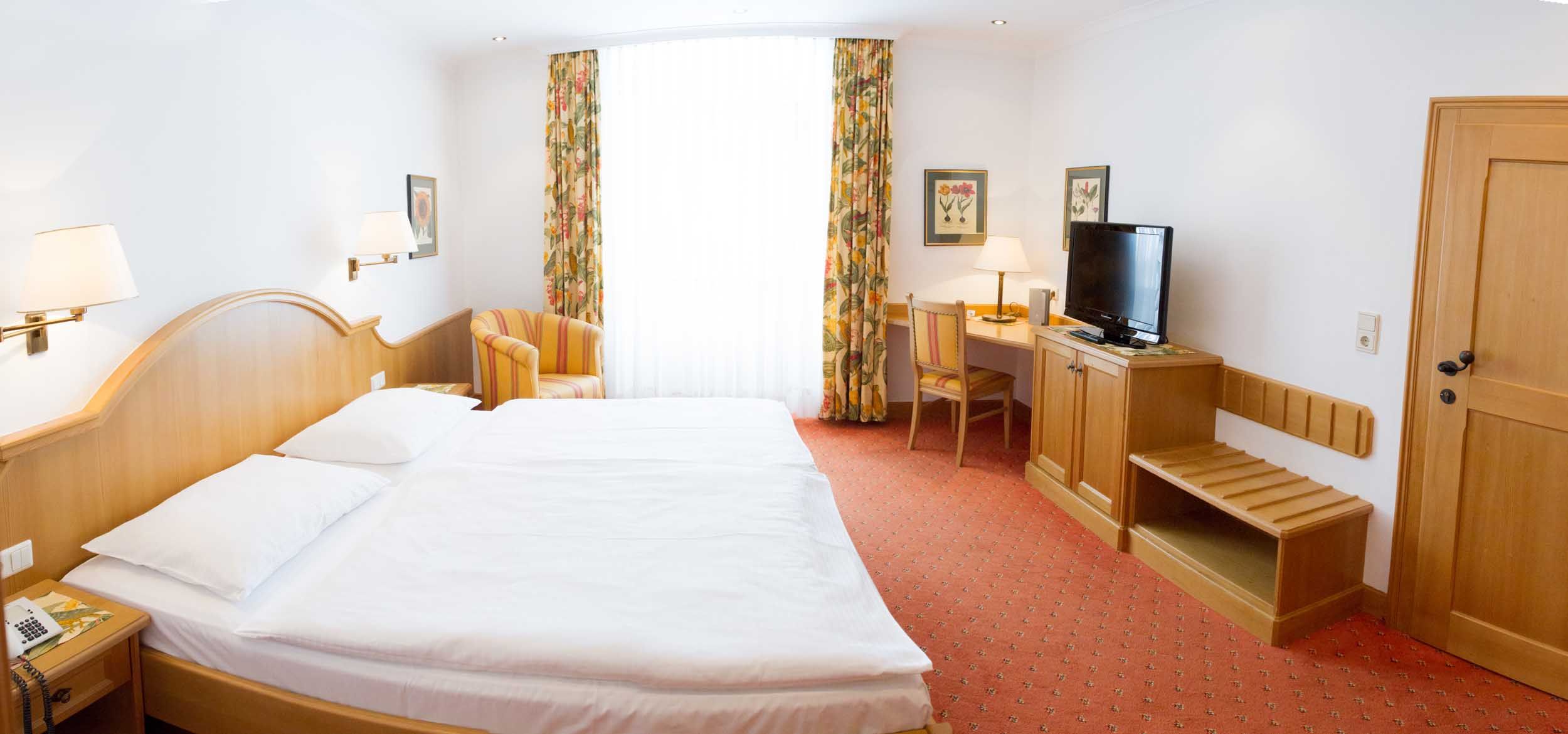 Ein Hotelzimmer mit Doppelbett, Schreibtisch, Fernseher und Sessel. Helle Vorhänge und florale Vorhänge schmücken das Fenster.