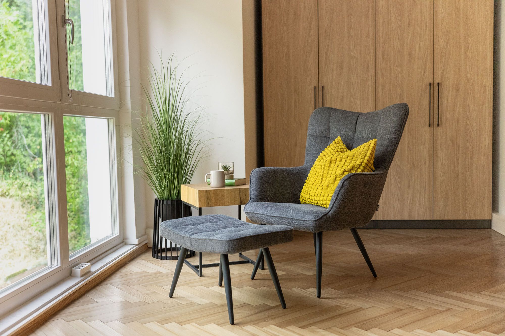 Gemütliche Sitzecke in der Ferienwohnung Turmblick mit grauem Sessel, gelbem Kissen, Hocker, Beistelltisch, Pflanze und großem Fenster.