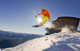 Ein Skifahrer springt vor dem Terzerhaus auf der Gemeindealpe in die Luft, umgeben von schneebedeckten Bergen und blauem Himmel.