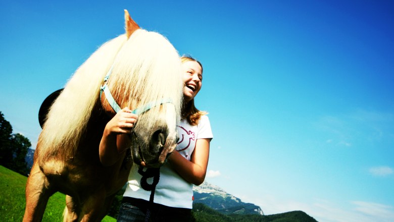 A girl laughs next to a horse in a green meadow with a blue sky in the background.