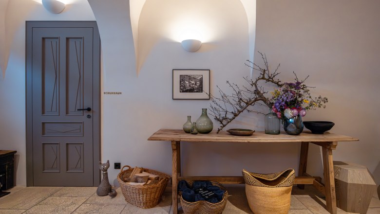 Innenraum mit Holztisch, Vasen, Blumen und Körben. Tür mit Aufschrift 'Schuhraum'.