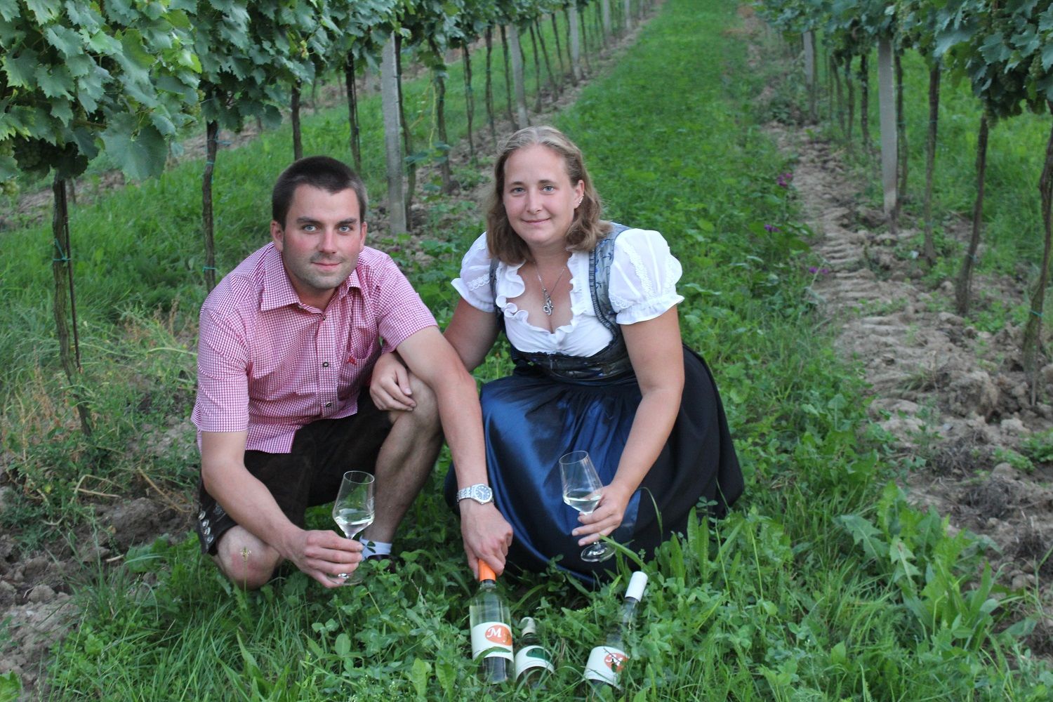 Zwei Personen sitzen in einem Weinberg mit Weingläsern und Flaschen.