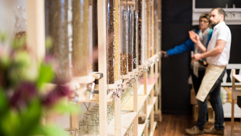 Innenansicht eines Unverpackt-Ladens mit Spendern für lose Lebensmittel und zwei Personen im Hintergrund.