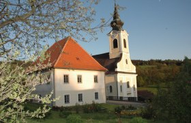 Kirche mit Pfarrhof in l&auml;ndlicher Umgebung, umgeben von B&auml;umen und Wiesen.