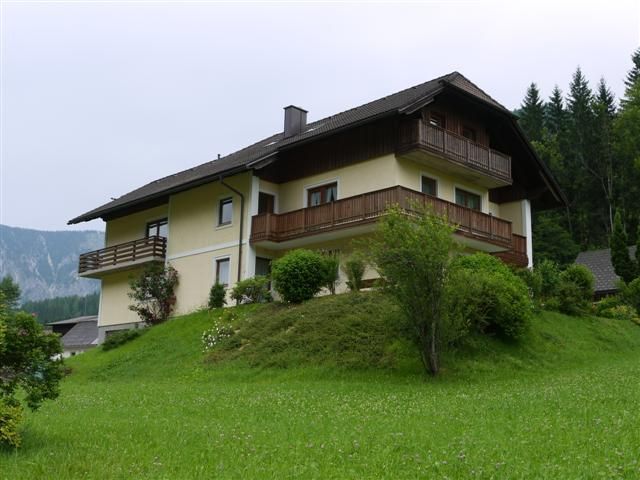 Ein zweistöckiges Haus mit Holzbalkonen auf einem grünen Hügel, umgeben von Bäumen und Wiesen.