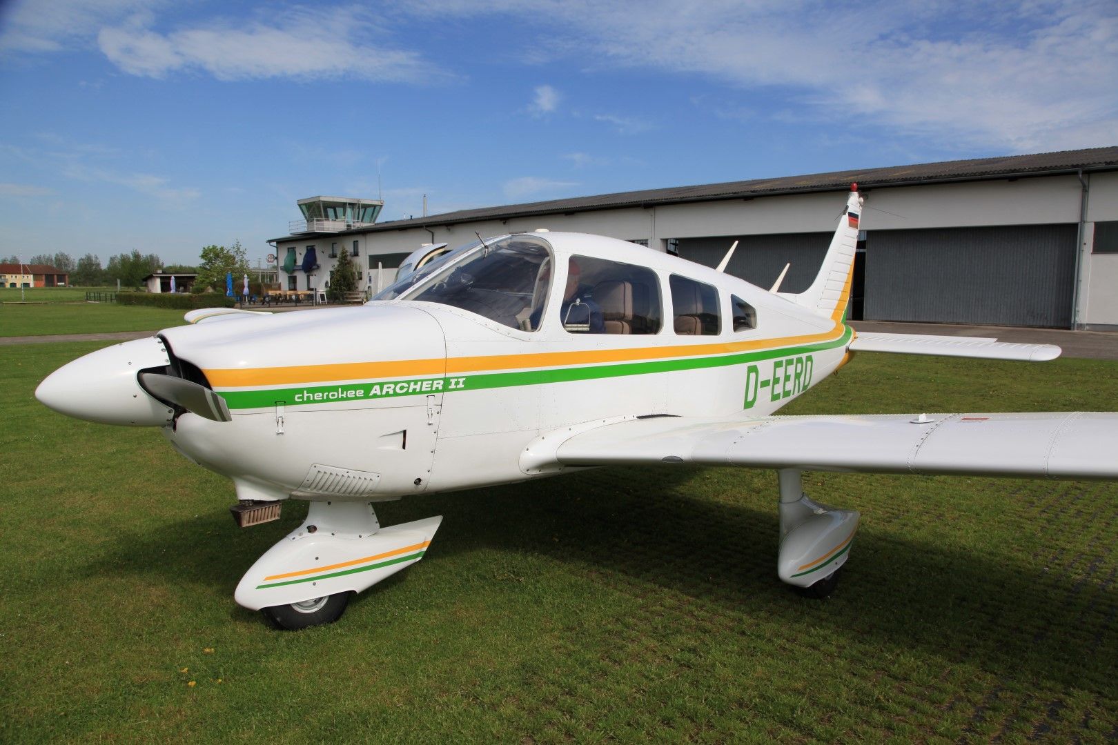 Ein kleines Flugzeug, Piper Cherokee Archer II, steht auf einer grünen Wiese vor einem Hangar und einem Kontrollturm.