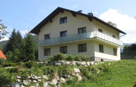 Ein zweist&ouml;ckiges Haus mit Balkon in einer gr&uuml;nen, h&uuml;geligen Landschaft.