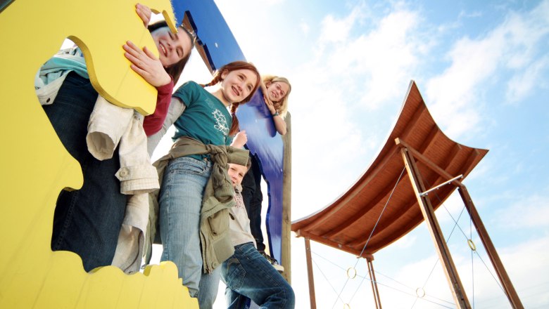 Kinder spielen auf einem bunten Klettergerüst mit einem Holzdach im Hintergrund.
