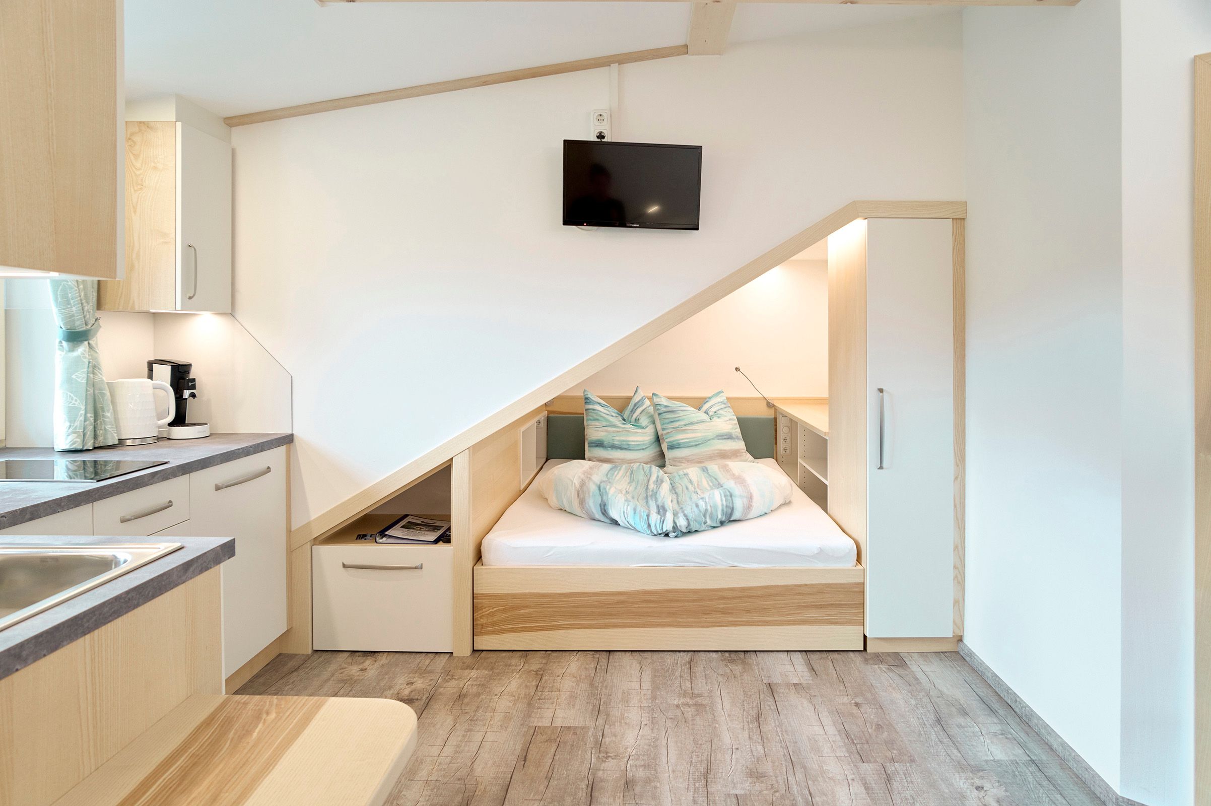 Kompaktes Apartment mit eingebautem Bett unter einer Dachschräge, Küchenzeile und Fernseher.
