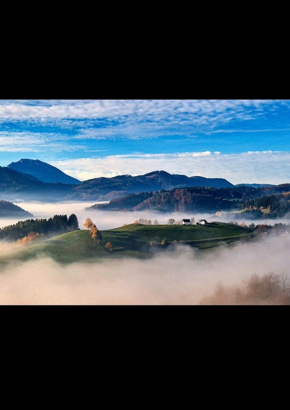 Eine malerische Landschaft mit grünen Hügeln, umgeben von Nebel und Bergen im Hintergrund unter einem blauen Himmel.
