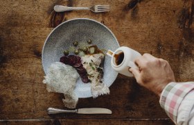 A plate of fish, beet and capers on a wooden table. A hand pours sauce from a jug over it.