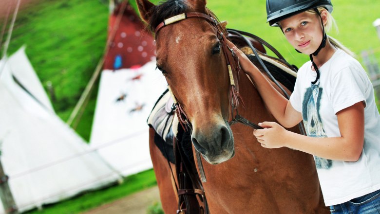 Ein Mädchen mit Helm steht neben einem braunen Pferd auf einem Bauernhof.